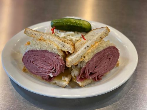 A plate with a sliced sandwich filled with corned beef and sauerkraut, accompanied by a pickle and coleslaw.