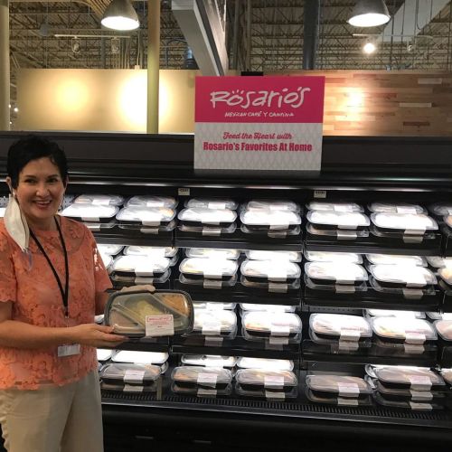 A woman holds a food container in front of a display of pre-packaged meals at a grocery store.