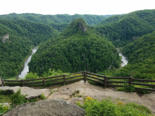 A scenic view of lush green mountains and a winding river, framed by a wooden railing in the foreground.