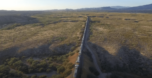 Aerial view of a long train traveling through a vast, arid landscape with mountains in the background.