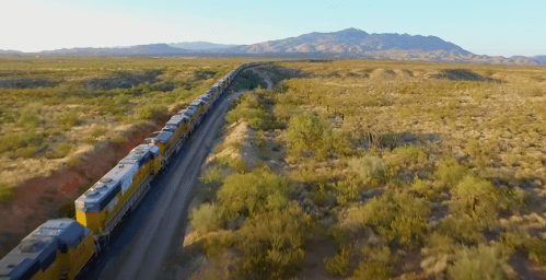 A long freight train travels through a desert landscape with mountains in the background under a clear blue sky.