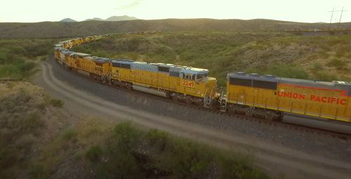 A Union Pacific freight train winds through a mountainous landscape at sunset, surrounded by greenery.