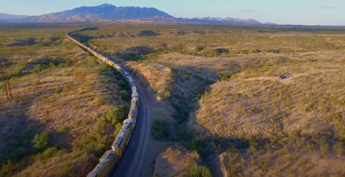 Aerial view of a winding train traveling through a vast, arid landscape with mountains in the background.