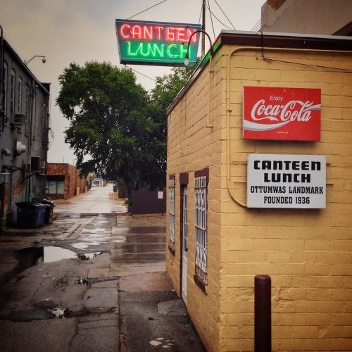 A yellow building with "Canteen Lunch" signs and a neon "Canteen Lunch" sign above, in a rainy alleyway.