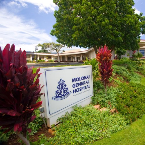 Sign for Moloka'i General Hospital surrounded by greenery and colorful plants under a clear blue sky.