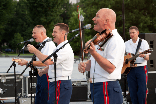 Four musicians in military uniforms perform on stage, singing and playing instruments at an outdoor event.