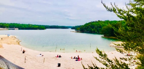 A serene lake surrounded by trees, with a sandy beach and people enjoying the water on a cloudy day.