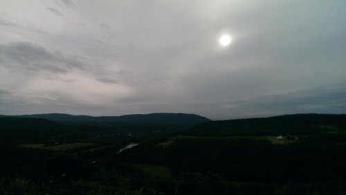 A cloudy sky over rolling hills and a river, with the sun partially visible in the background.