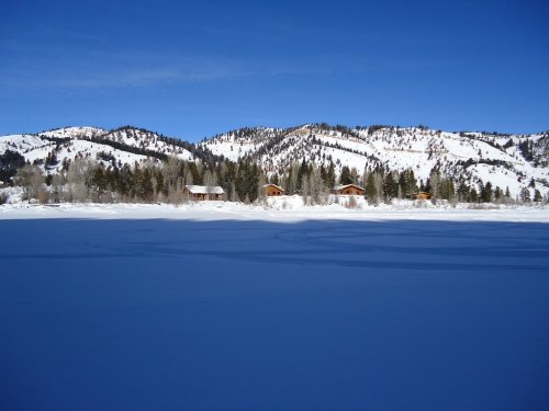 Snow-covered landscape with a frozen lake, surrounded by mountains and a few wooden cabins under a clear blue sky.