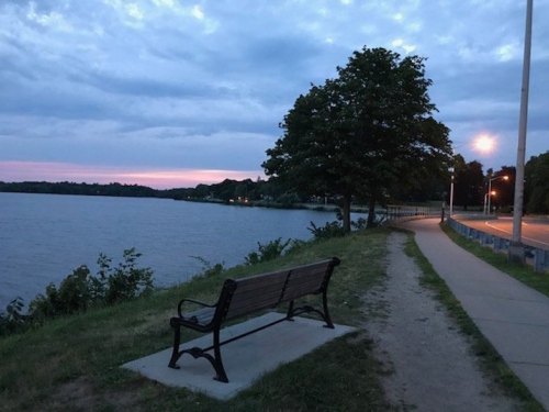 A quiet lakeside path at dusk, featuring a bench and trees, with soft lighting along the walkway.