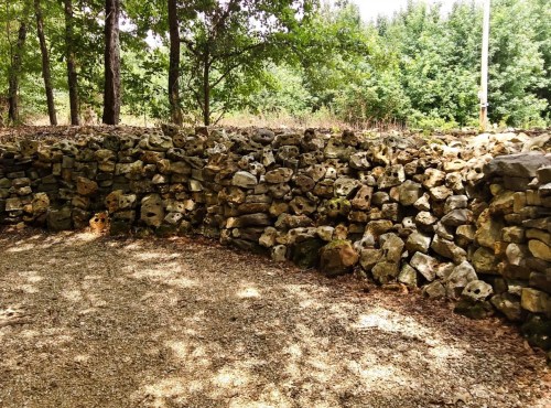 A stone wall made of irregular rocks, surrounded by trees and a gravel path in a natural setting.