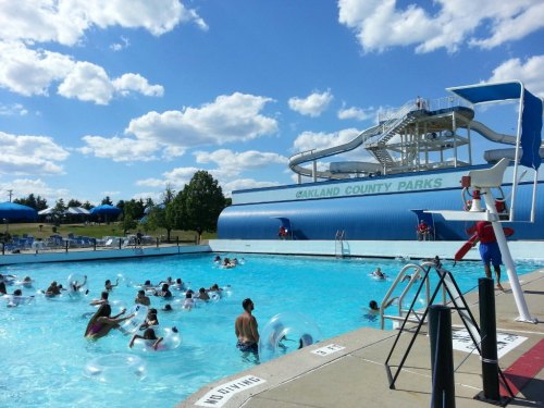 A busy outdoor pool with people swimming and playing, featuring a large water slide and lifeguard on duty.