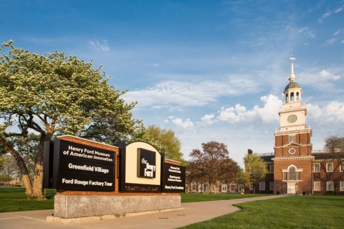 Sign for the Henry Ford Museum of American Innovation with the museum building and trees in the background under a blue sky.