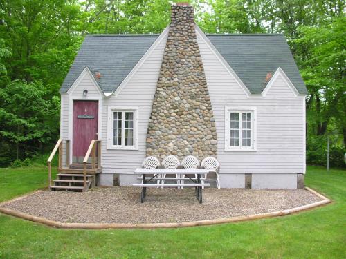 A small, charming house with a stone chimney, surrounded by greenery and featuring a picnic table in front.