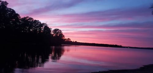 A serene lake at sunset, with vibrant pink and purple skies reflecting on the calm water, framed by silhouettes of trees.