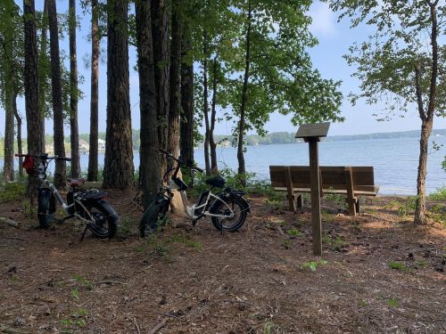 Two bicycles parked near a serene lake, surrounded by trees, with a bench and a signpost nearby.