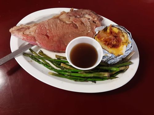 A plate with a large slice of roast beef, baked potato, asparagus, and a small cup of gravy.