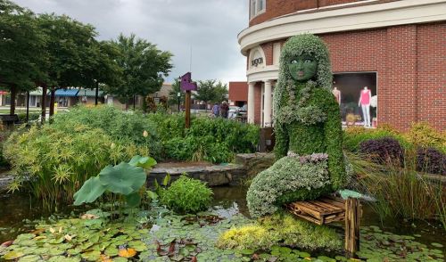 A large, green sculpture of a seated woman made of plants, surrounded by a pond with lily pads and flowers.