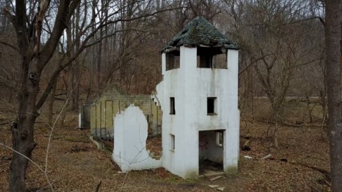 An abandoned, partially collapsed building with a tower, surrounded by bare trees and overgrown foliage.