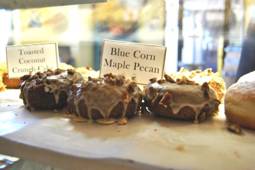 Two donuts topped with maple glaze and pecans, labeled "Blue Corn Maple Pecan," displayed alongside a coconut crunch cake.