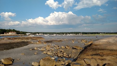 A scenic beach with rocky formations, boats in the distance, and fluffy clouds in a blue sky.