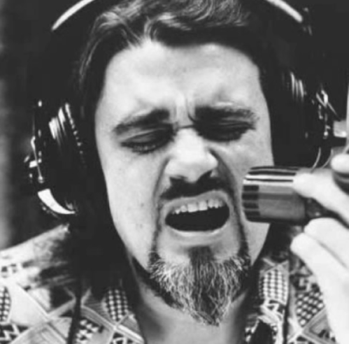 A man with long hair and a beard passionately sings into a microphone while wearing headphones.