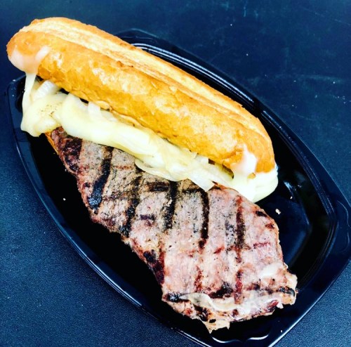 A grilled steak sandwich with melted cheese and sautéed onions in a toasted bun on a black plate.