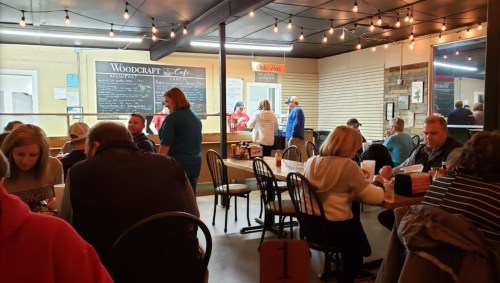 A busy café interior with patrons dining, staff serving, and a menu board in the background. Warm lighting and a cozy atmosphere.