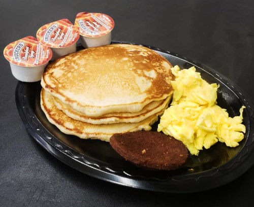 A black plate with three pancakes, scrambled eggs, and a chocolate cookie, accompanied by syrup cups.