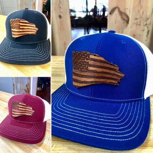 Three trucker hats in navy, blue, and burgundy, each featuring a wooden American flag design on the front.