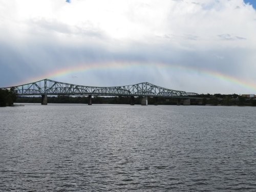 A bridge spans a river under a cloudy sky, with a faint rainbow arching above.