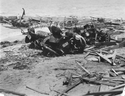 Wreckage of old cars and debris scattered on a beach, with water in the background.