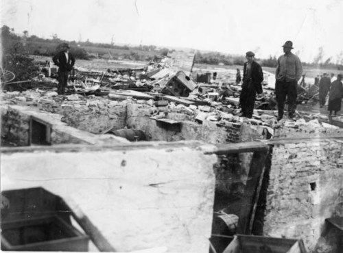 Three men stand on the ruins of a collapsed building, surveying the destruction around them.