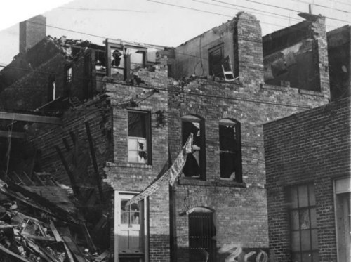 Black and white photo of a damaged brick building with a collapsed roof and debris scattered around.