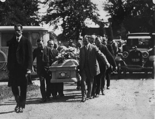 A black-and-white photo of a funeral procession with men carrying a casket, surrounded by vintage cars.