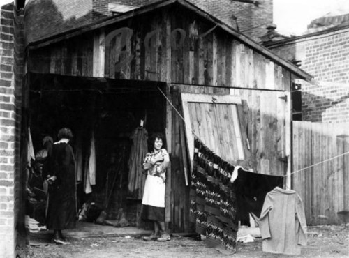 A black-and-white photo of a wooden shed with women outside, hanging clothes on a line.