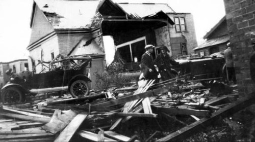 A damaged house with a collapsed front, surrounded by debris and an old car, with people inspecting the scene.
