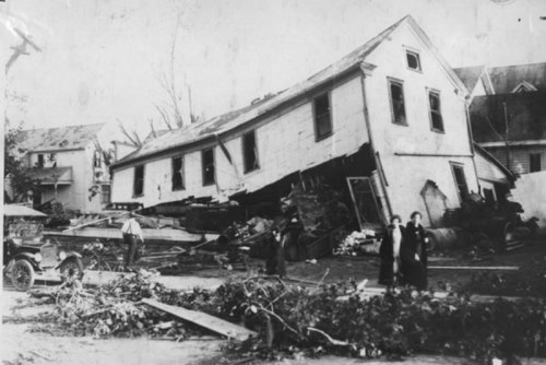 Historic photo of a collapsed house with debris, two women in black dresses, and a vintage car nearby.
