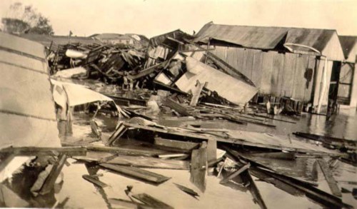 A black-and-white photo of a flooded area with damaged buildings and debris scattered around.