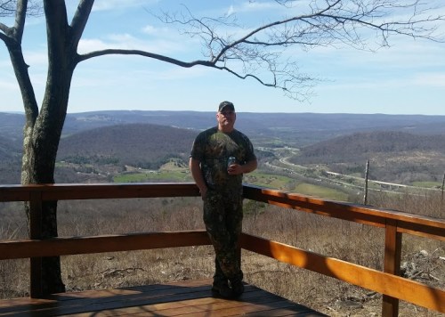 A man in camouflage stands on a wooden deck overlooking a scenic valley with rolling hills and a clear blue sky.