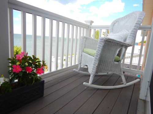 A white rocking chair on a porch overlooking the water, with colorful flowers in a planter nearby.