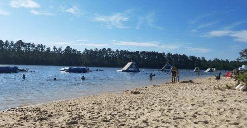 A sandy beach by a lake with people swimming and inflatable structures in the water under a clear blue sky.