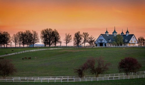 A picturesque farm at sunset, featuring a white barn with spires and grazing horses on a green hillside.