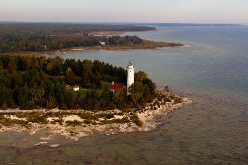 Aerial view of a lighthouse on a rocky shore, surrounded by trees and water, with a calm horizon in the background.
