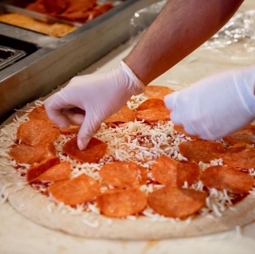 A hand in a glove places pepperoni slices on a cheese-covered pizza crust before baking.