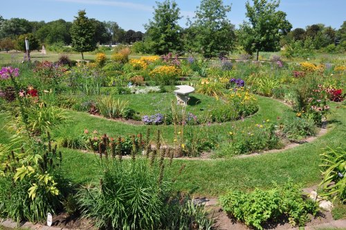 A vibrant garden with colorful flowers arranged in a spiral pattern, surrounded by lush greenery and trees.