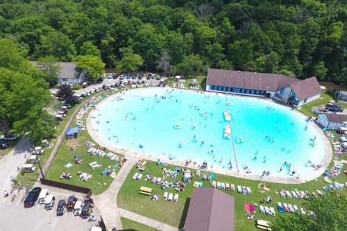 Aerial view of a large swimming pool surrounded by greenery, with people swimming and lounging on deck chairs.
