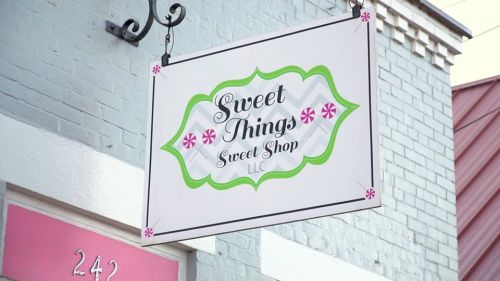 Sign for "Sweet Things Sweet Shop" with decorative elements, hanging from a building. Address: 242.