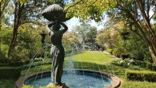 A statue of a woman holding a pot stands by a fountain in a lush garden with greenery and flowers.