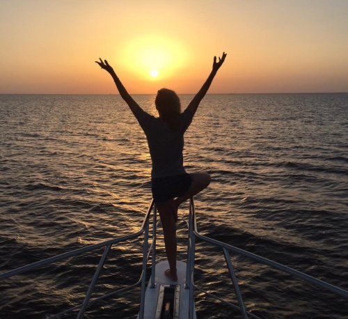 Silhouette of a person on a boat at sunset, arms raised, with a calm sea in the background.
